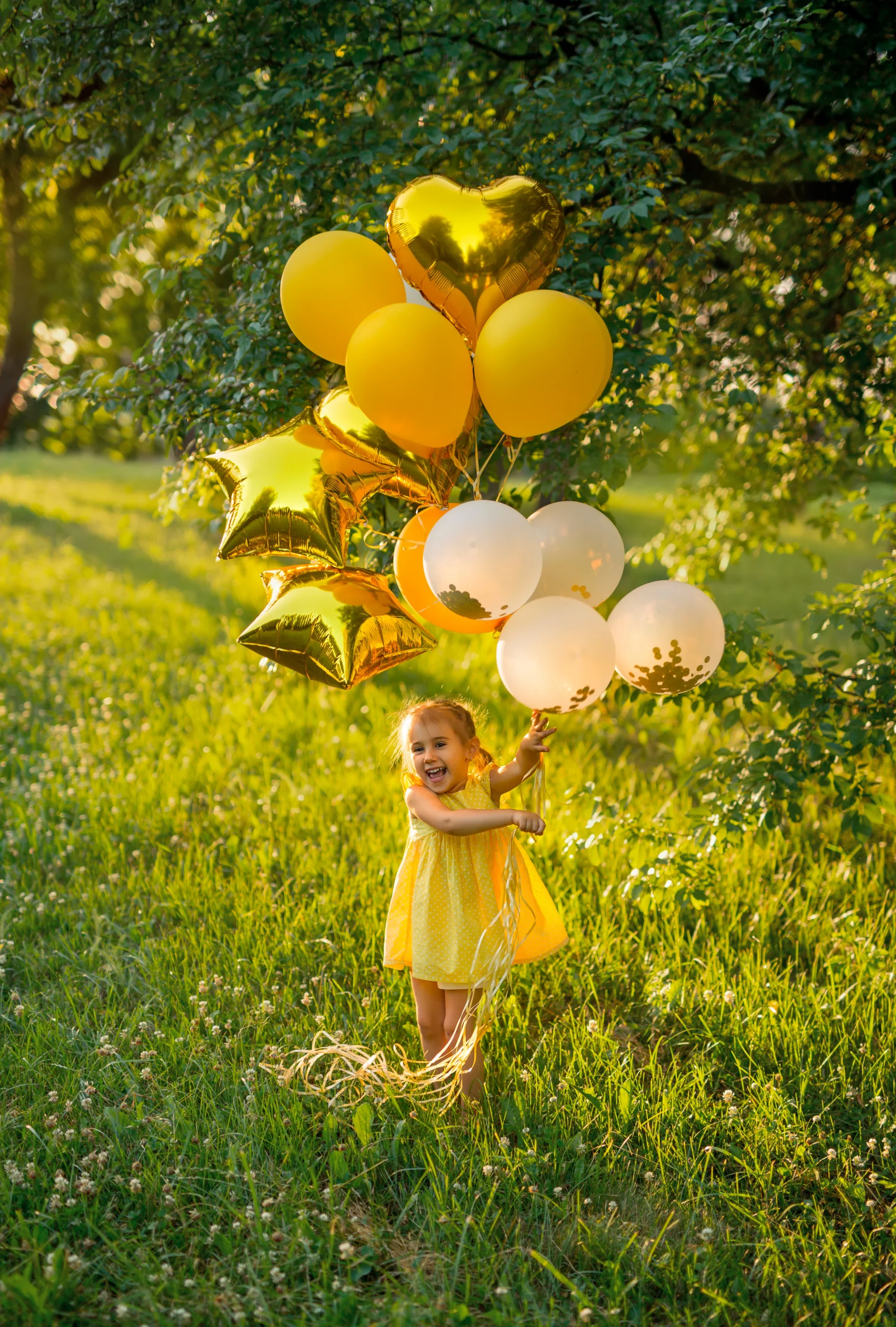 Photo d'une petite fille en robe jaune qui tient plusieurs ballons jaunes et blancs et qui court dans l'herbe